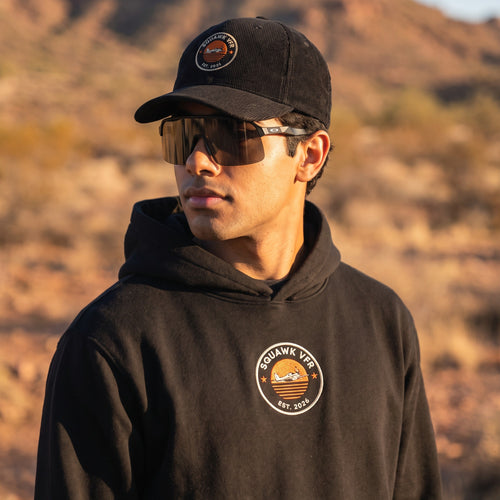 On-model — SquawkVFR Embroidered Roundel Hoodie in vintage black with matching cap, worn at golden hour, SquawkVFR